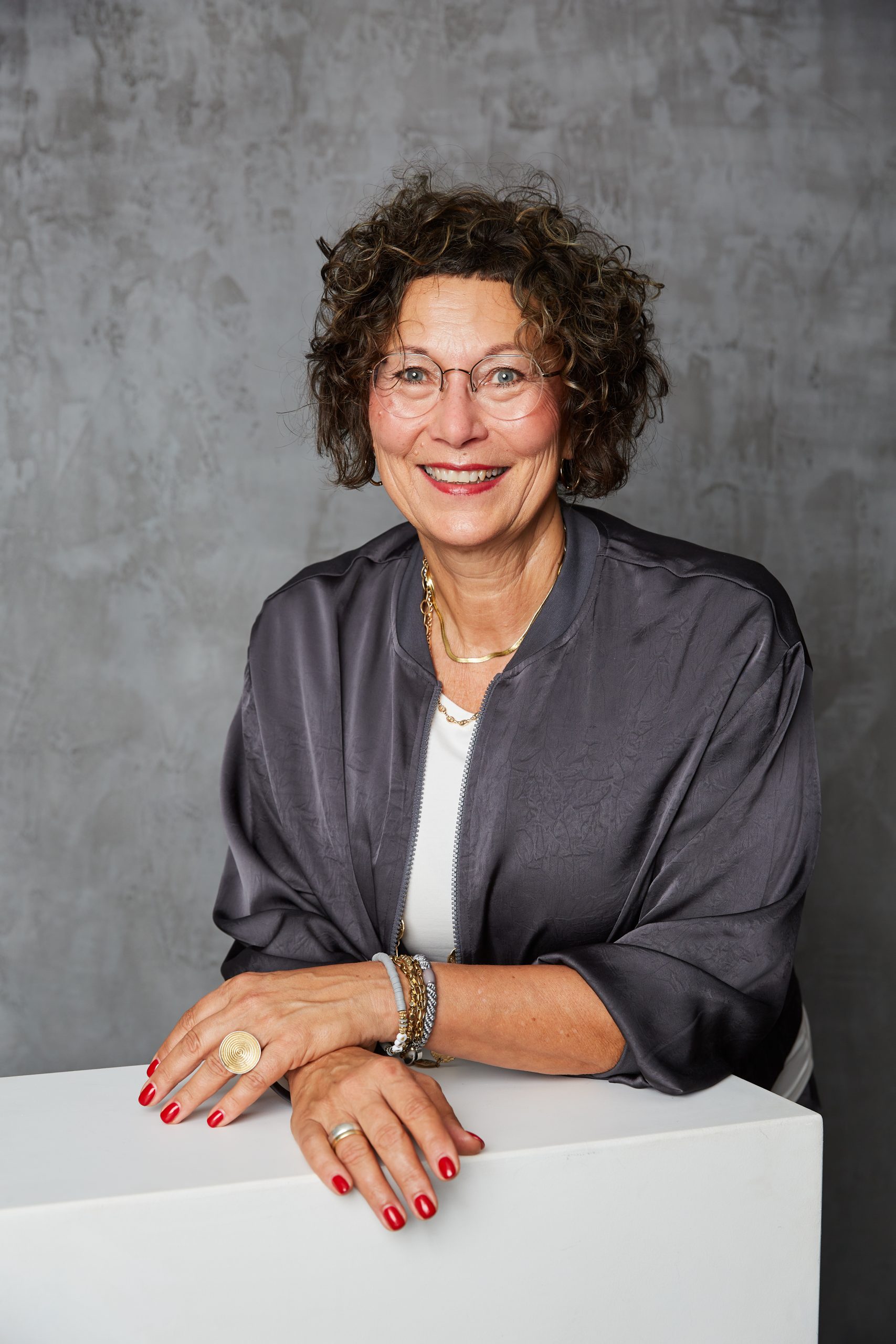 Portrait - Birgit Schneider Trauerrednerin Birgit Schneider lächelt in die Kamera und lehnt sich leicht auf ein weißes Pult. Sie trägt ein graue Bluse.